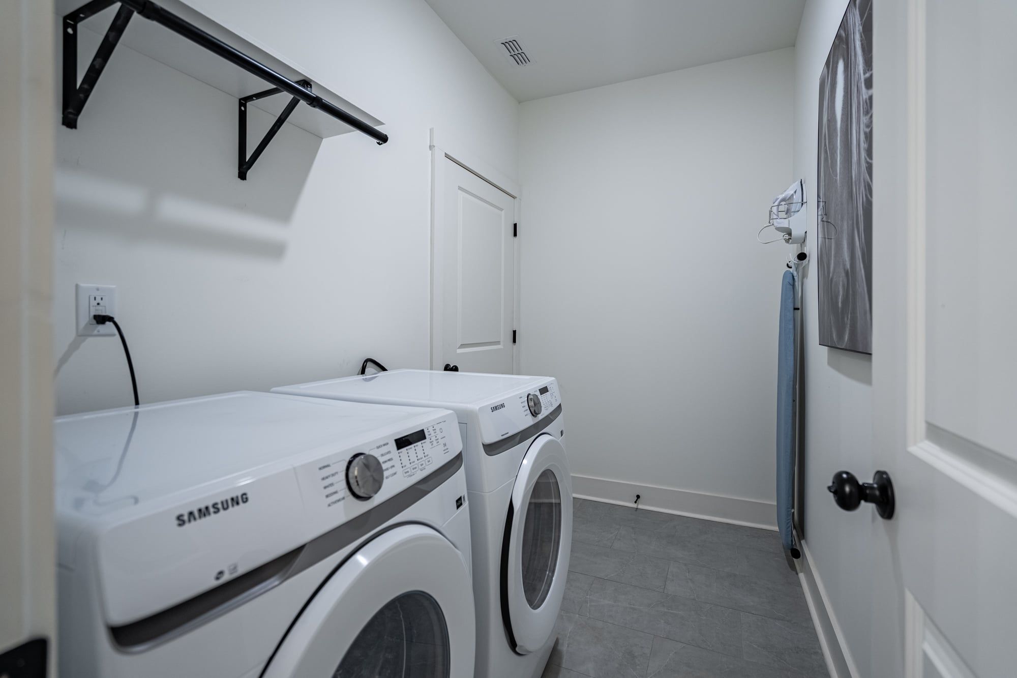 A laundry room with two Samsung washing machines, a drying rack, and a door with a towel rack.