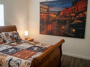 Ein Schlafzimmer mit einem Bett, das eine Western-Steppdecke zeigt, einem Nachttisch mit Lampe und einem großen Poster einer Westernstadt an der Wand.