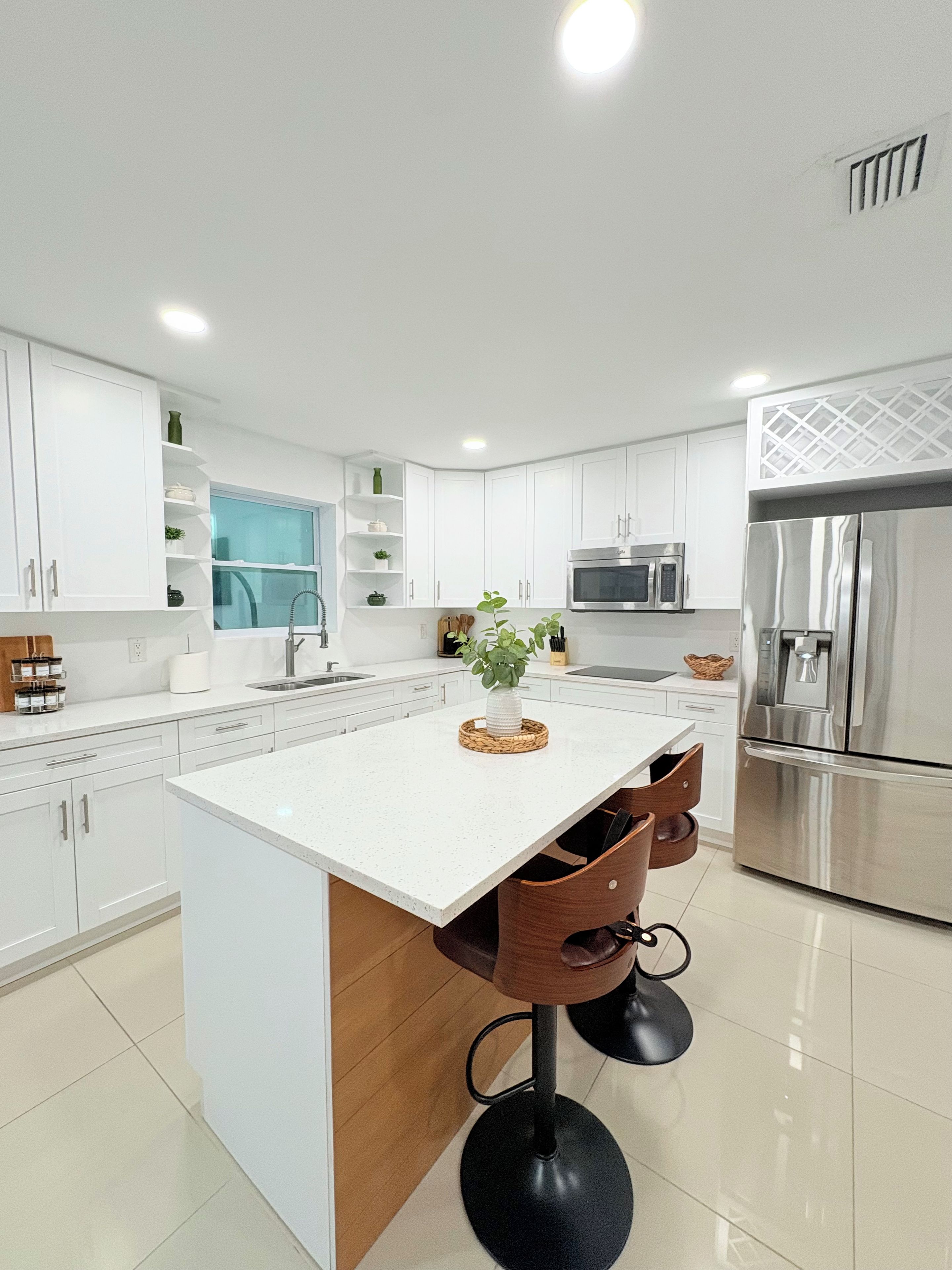 A modern kitchen with white cabinetry, a central island with stools, a stainless steel refrigerator, and a microwave.