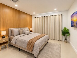 A modern bedroom with a wooden panel wall, a bed with multiple pillows, a nightstand with a lamp, a large window with curtains, and a potted plant. - 3-bedroom house in amazing Lake Worth Beach with AC comfort (Lake Worth Beach)
