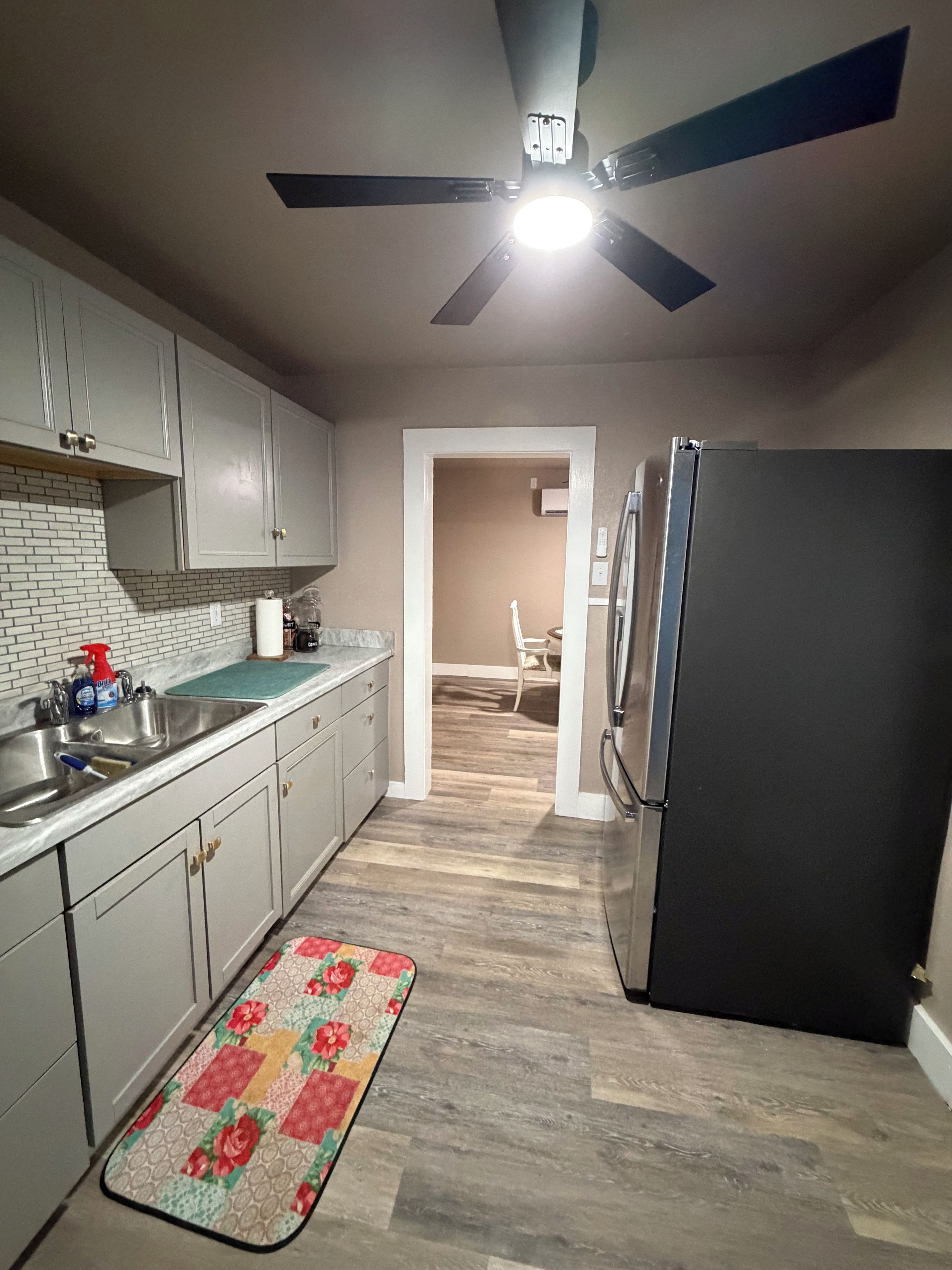A kitchen with a black refrigerator, white cabinets, a wooden floor, and a ceiling fan.