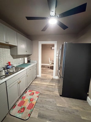 Una cocina con un refrigerador negro, armarios blancos, piso de madera y un ventilador de techo.