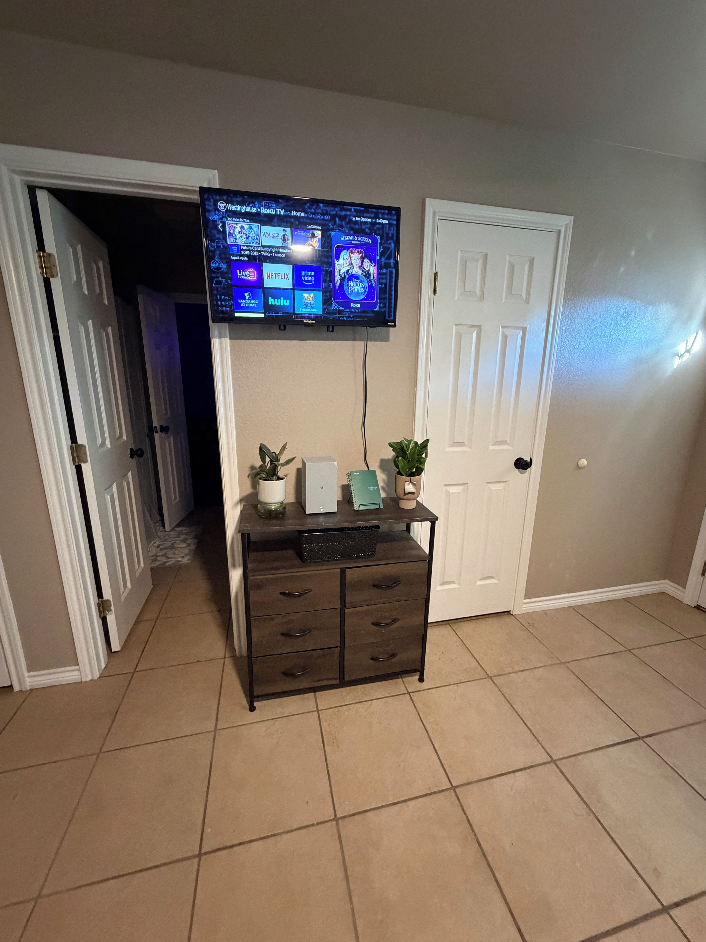 Een kamer met een flatscreen televisie aan de muur, een donkere houten kast met lades, en een betegelde vloer.