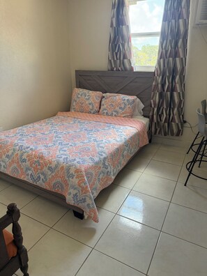 A neatly made bed with patterned bedding, a wooden headboard, and a window with curtains. - Cozy 1-bedroom studio  apartment near  Bridgetown with AC Opp Sea, Beach, bars! (Bridgetown)
