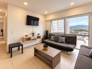 A modern living room with a sofa, a coffee table, a TV mounted on the wall, and a window with a city view. - New Listing (Zapopan)