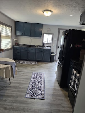 Una cocina compacta con armarios oscuros, un refrigerador negro y una alfombra estampada sobre piso de madera.