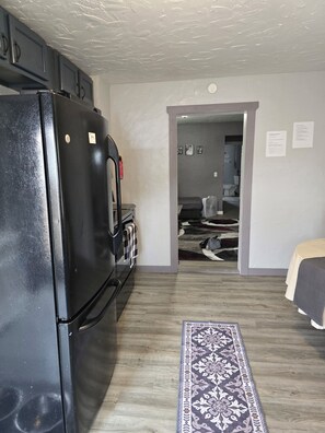 A compact kitchen with a refrigerator, a stove, and a microwave. There is a patterned rug on the floor, and a bed is visible in the adjacent room.