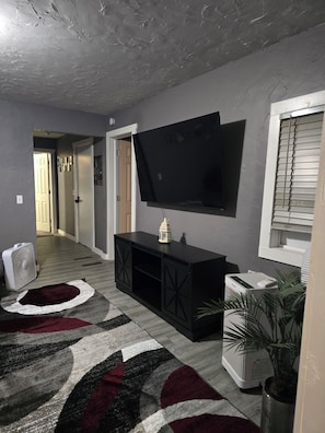 Una sala de estar con un televisor de pantalla plana montado en la pared, un mueble de entretenimiento negro, un radiador blanco y una alfombra estampada.