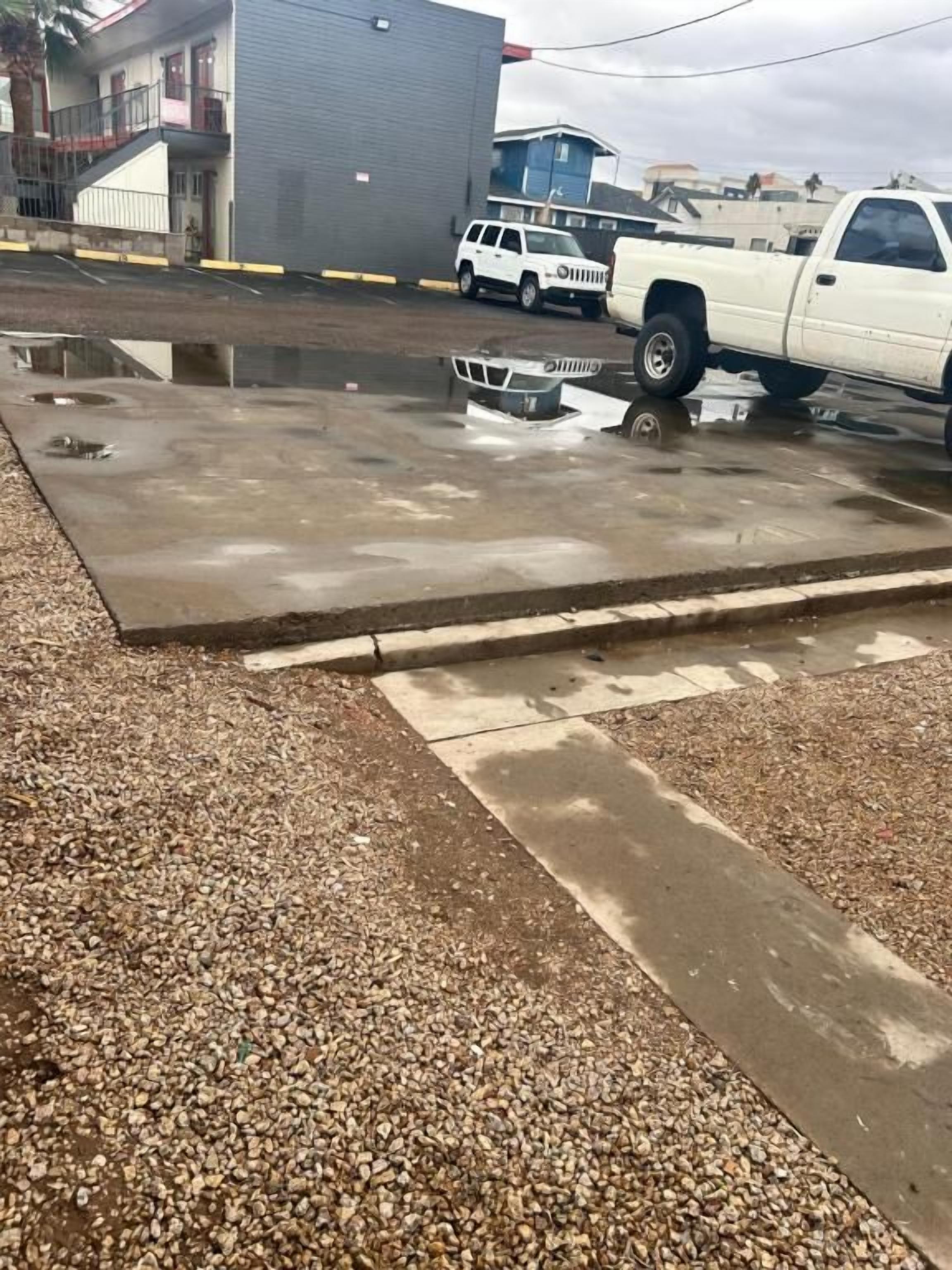 A parking area with a white pickup truck parked on a wet surface, surrounded by gravel and a concrete barrier.