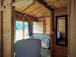 Interior de una cabaña de madera con sofá, una silla y una puerta que da al exterior.