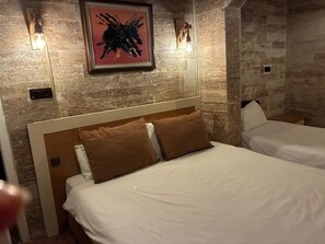 Habitació d'hotel amb paret de pedra, un quadre abstracte emmarcat i dos llits amb llençols blancs i almoadins marrons.