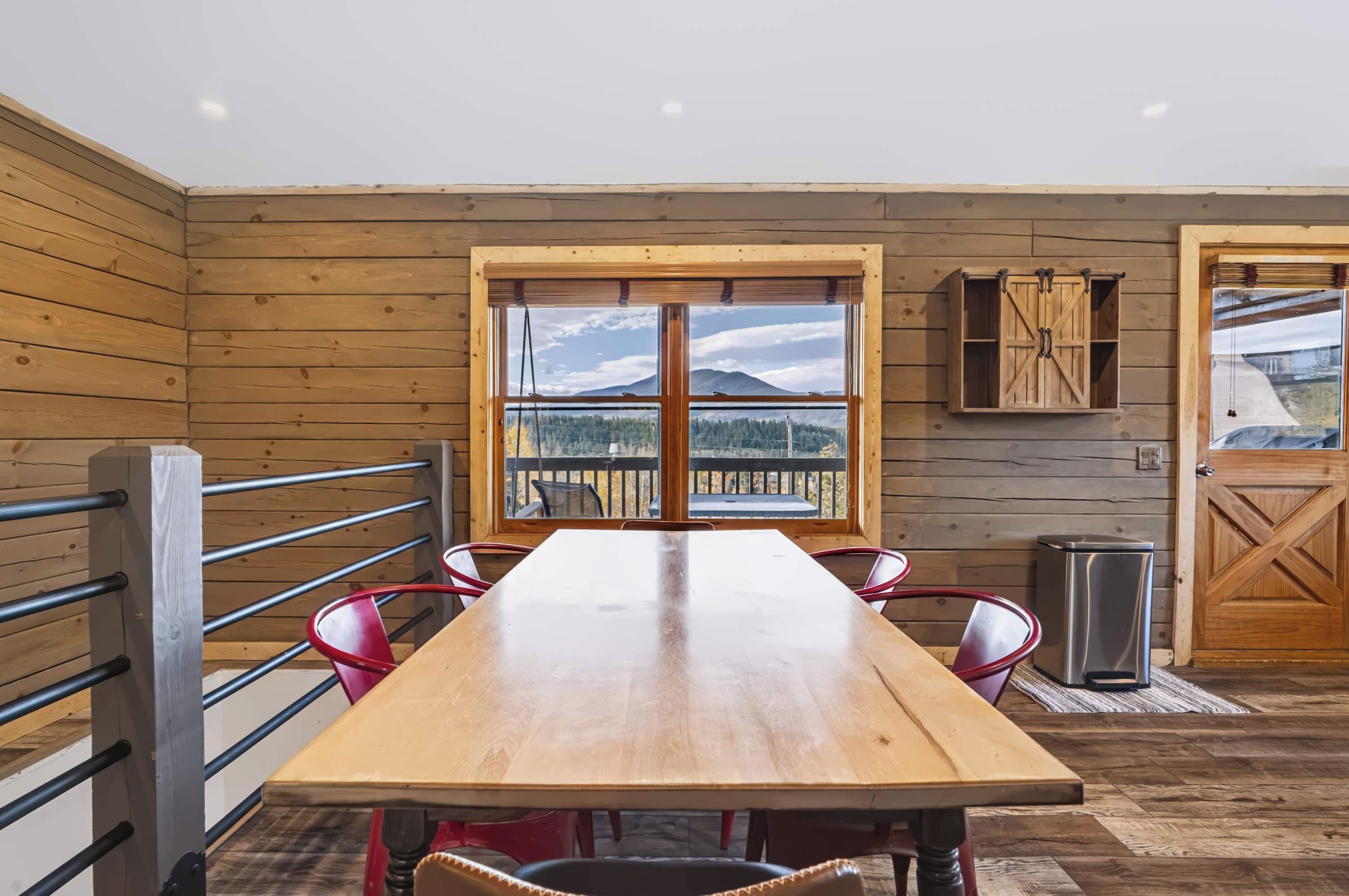 A wooden interior with a long table, chairs, a window with a view, and a door.