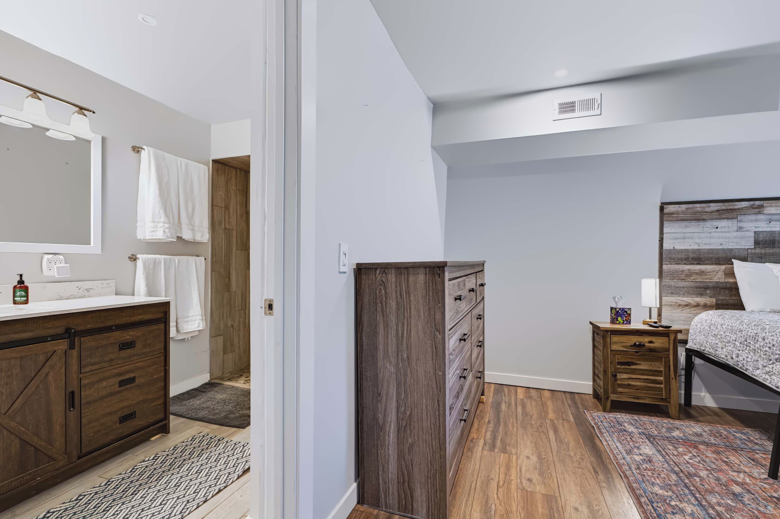 A modern bedroom with a wooden dresser, a bed with a headboard, a nightstand, and a bathroom with a mirror and towel rack.