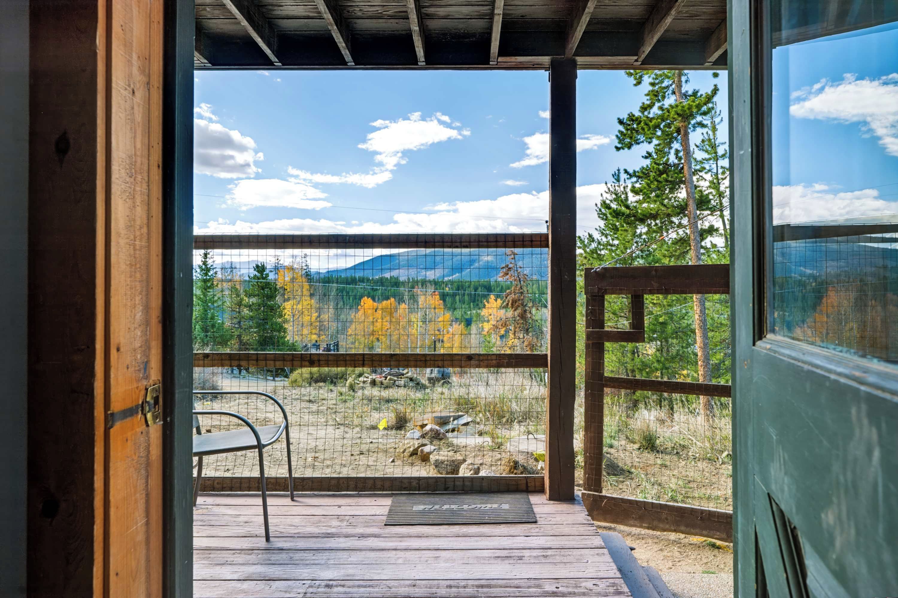 Una terraza de madera con una silla metálica, un felpudo y vistas a un paisaje montañoso con follaje otoñal.