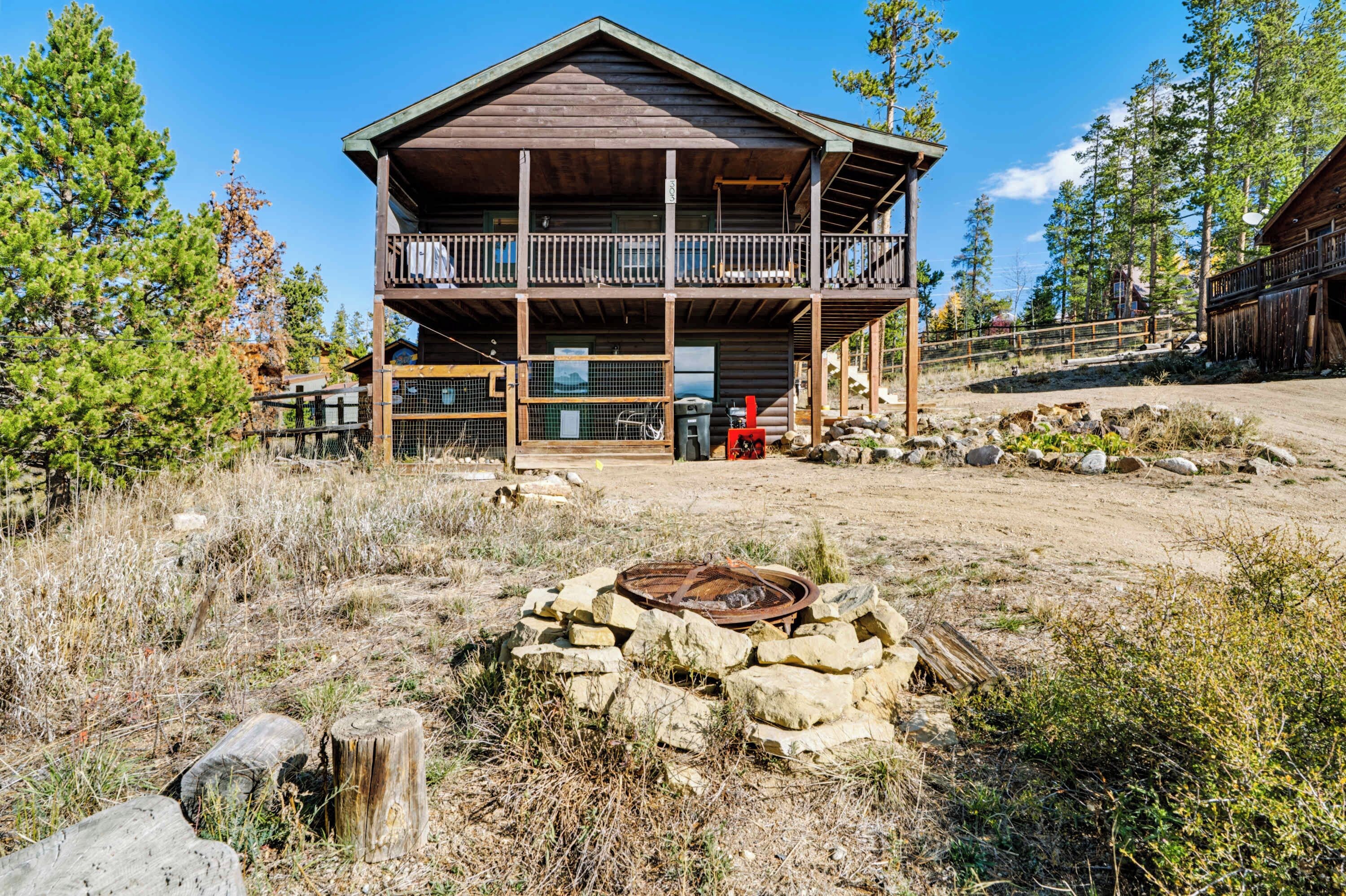 A two-story log cabin with a balcony, surrounded by a natural landscape with trees and a fire pit in the foreground.