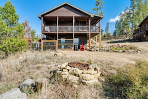A two-story log cabin with a balcony, surrounded by a natural landscape with trees and a fire pit in the foreground.