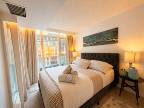 A hotel room with a large bed, a window with curtains, a wall-mounted artwork, and a bedside table with a lamp. - Luxury Skyline View | 2 Master Suites | CityPlace (Toronto)