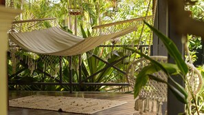 Una hamaca suspendida sobre una terraza de madera con un tapete tejido debajo, rodeada de exuberante vegetación tropical.