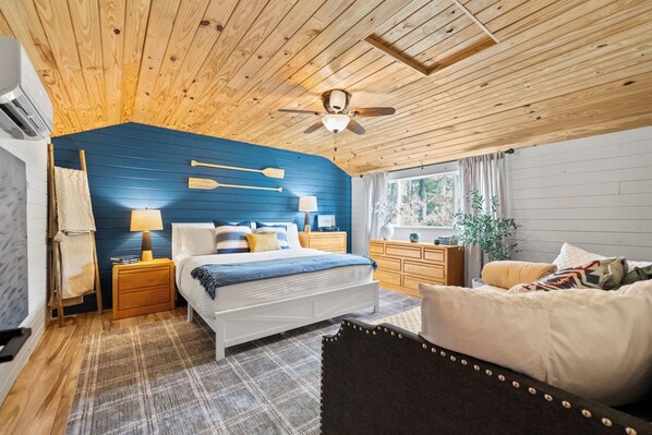A bedroom with a wooden ceiling, a bed with white bedding, a nightstand with a lamp, a dresser, and a window with curtains. - Reel Good Times on Mill Creek Waterfront Fishing & Family Retreat with Kayaks (Scottsboro)