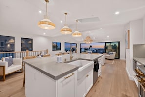 A modern kitchen with a central island, pendant lighting, and large windows offering a view of the cityscape. - Moonlight Dream - 4 Bedroom Panoramic Ocean Views (Encinitas)