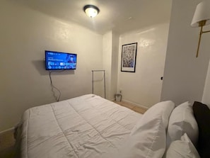 Un dormitorio con una cama, un televisor montado en la pared, un cuadro enmarcado y una lámpara de pie.