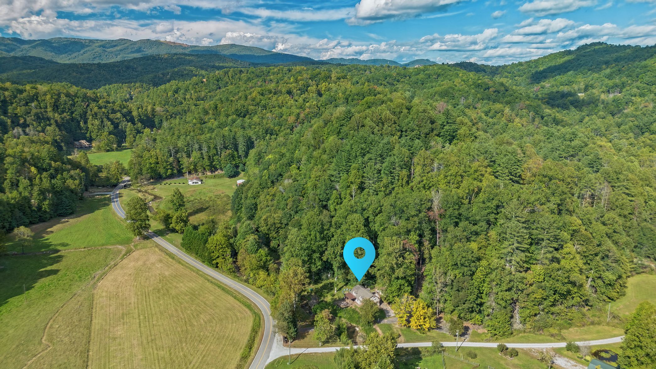An aerial view of a rural area with a winding road, a cluster of houses, and dense forested hills in the background.