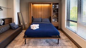 A modern bedroom with a bed, a sofa, a bedside table, a lamp, a fan, and a window with blinds. - Stylish Studio with Birmingham City Views D1 (Birmingham)