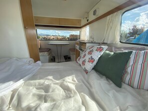 Interior compacto de una caravana con una cama, una mesita y una ventana que ofrece vistas al exterior.