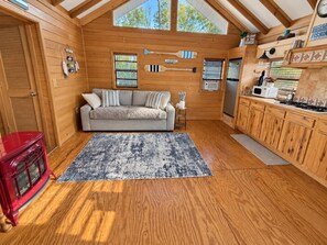 A cozy cabin interior with a wooden floor, a sofa, a red stove, and a kitchen area. - Peaceful Riverfront Cabin with Screened Porch & Fishing! (Live Oak)