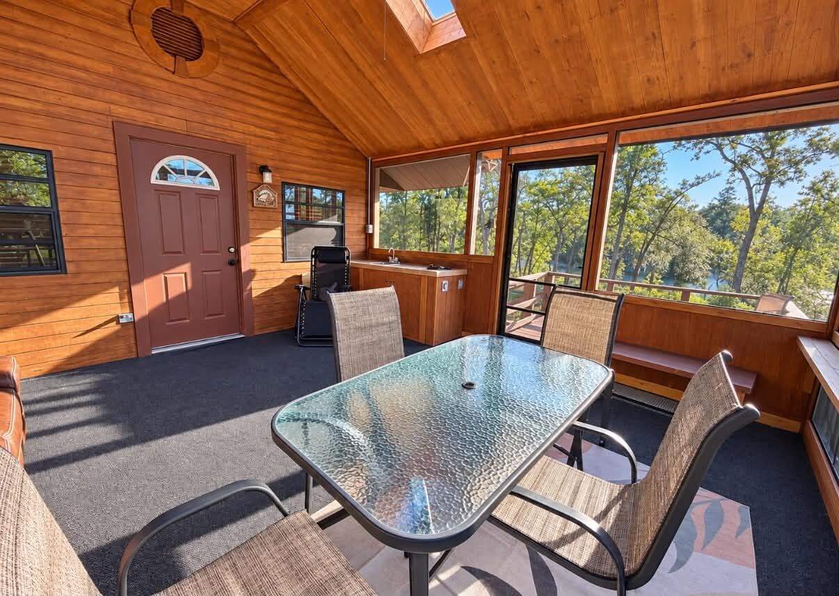 A wooden cabin with a glass table and chairs, a door, and large windows overlooking a forest.