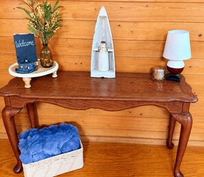 Une table en bois, un panneau d’accueil, une bouteille décorative, une lampe et un coussin bleu dans un panier.