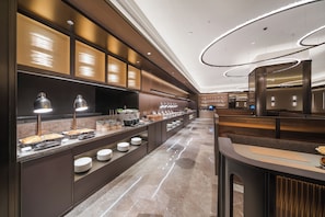 Um hall de hotel moderno com um balcão de recepção extenso, uma área de buffet com diversos alimentos e um espaço de estar com pé-alto e iluminação contemporânea.