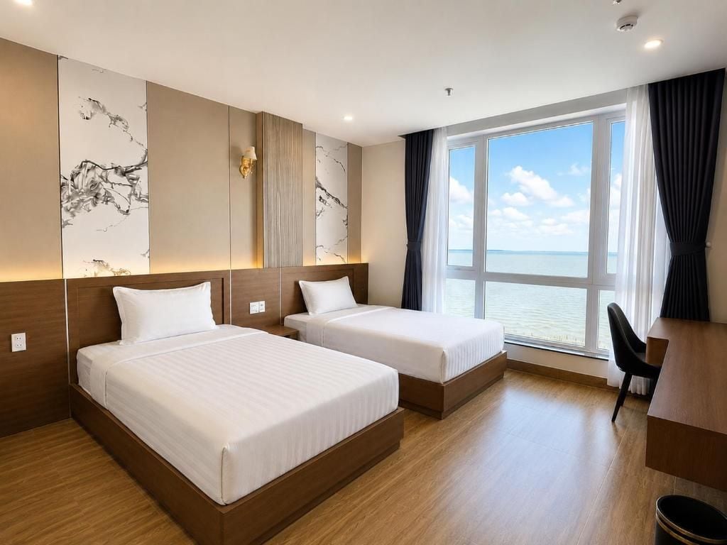 A hotel room with two beds, a desk, and a large window with a view of the sea.