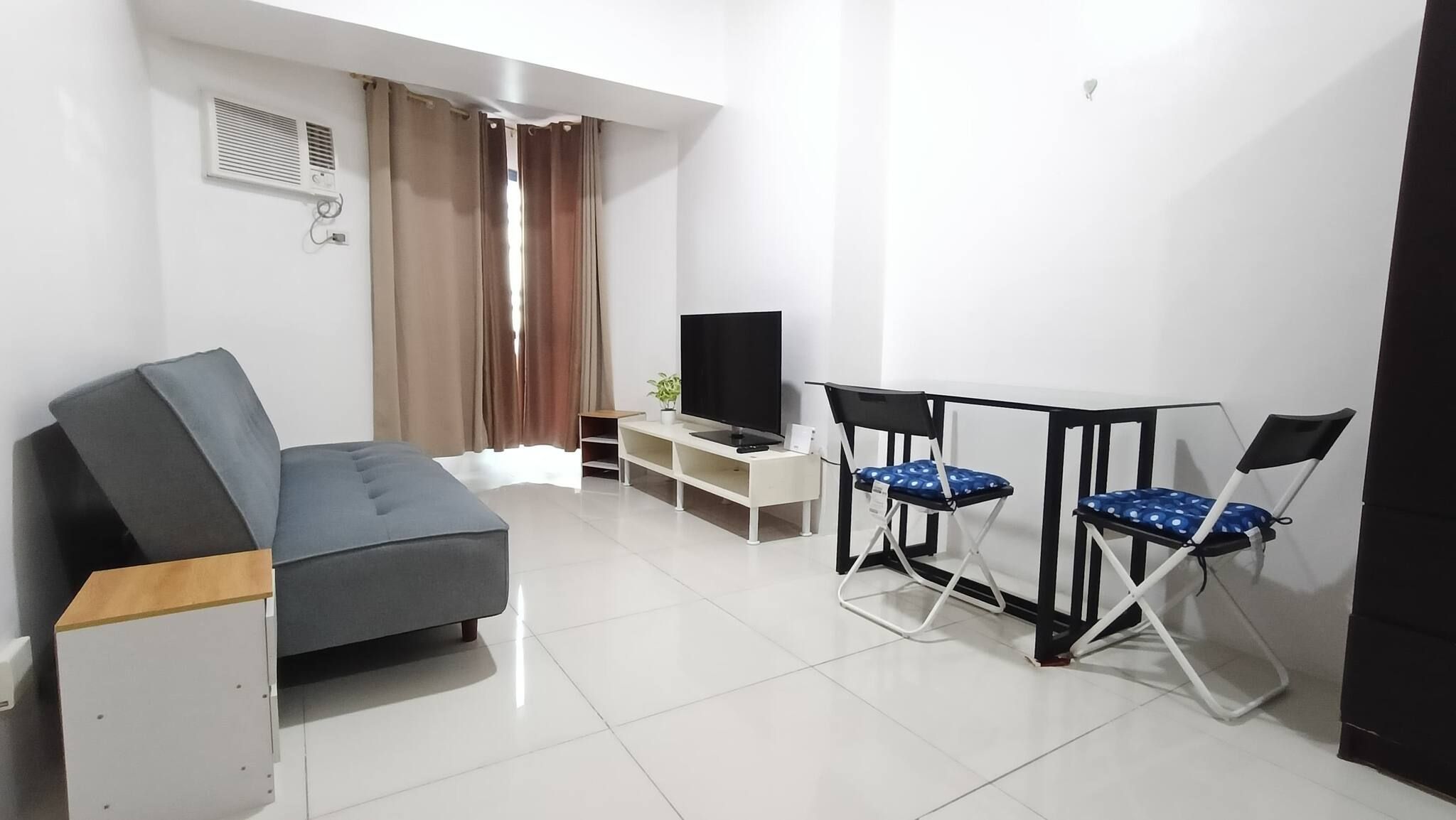 Una sala de estar moderna con un sofá, un televisor, una pequeña mesa con sillas y una ventana con cortinas.