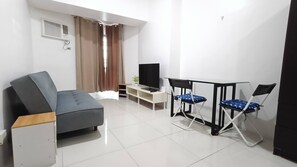 Una sala de estar moderna con un sofá, un televisor, una pequeña mesa con sillas y una ventana con cortinas.