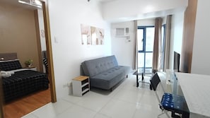 Ein modernes, helles Zimmer mit einem grauen Sofa, einem kleinen Tisch und einem Bett mit Kopfteil.