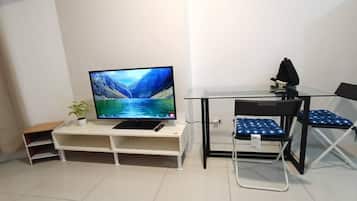 A compact living space with a flat-screen TV on a white stand, a small plant, a foldable table, and two chairs with blue cushions.