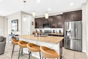 A modern kitchen with dark cabinets, a stainless steel refrigerator, a central island with bar stools, and pendant lighting. - Sunshine & Stay Awhile (Davenport)