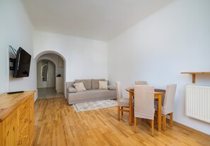 Een moderne woonkamer met een houten vloer, een grijze bank, een eettafel met stoelen en een aan de muur bevestigde televisie.