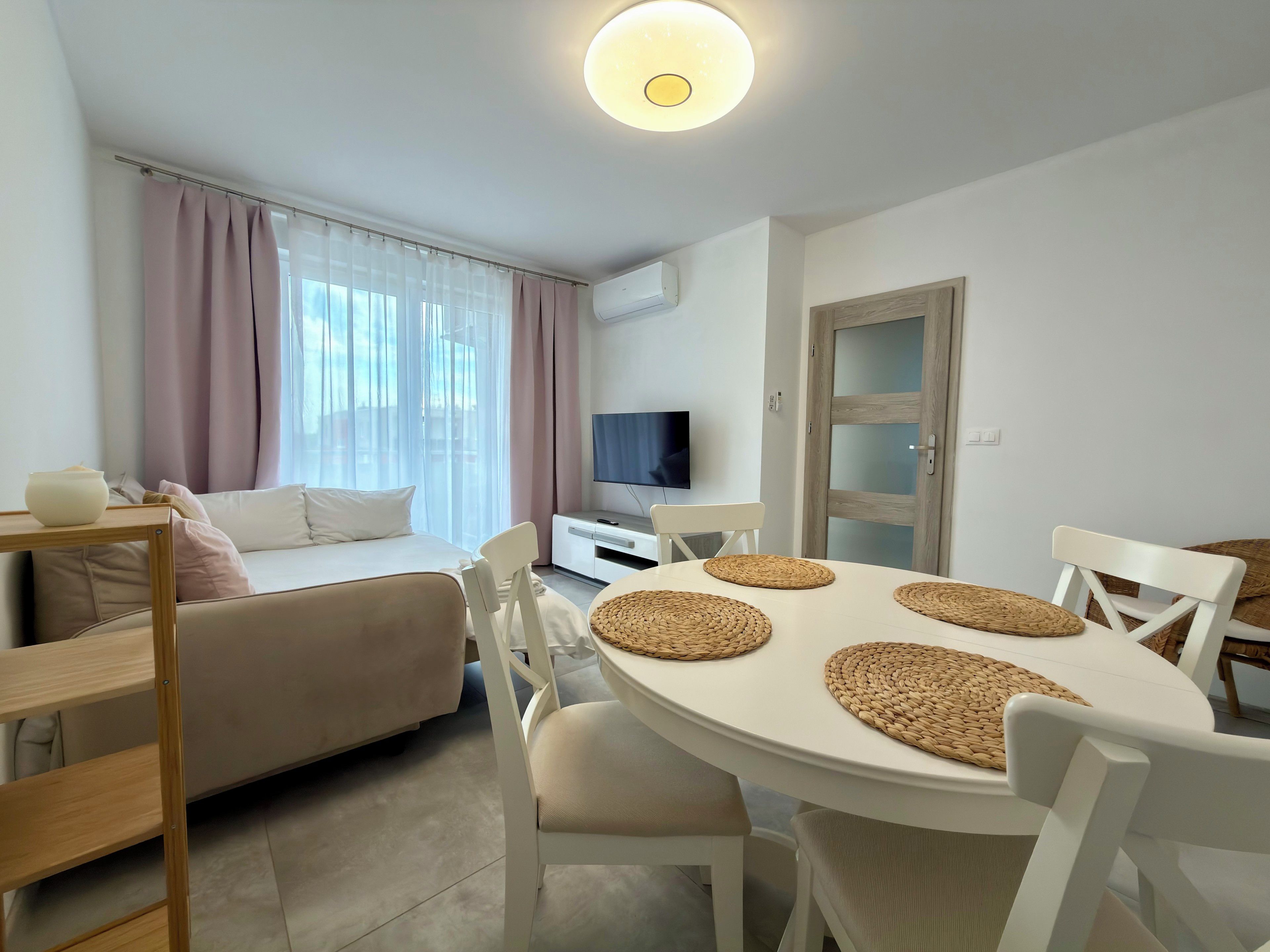 Une pièce moderne et bien éclairée, comprenant un lit, une table à manger avec des sets de table, un téléviseur et une porte.