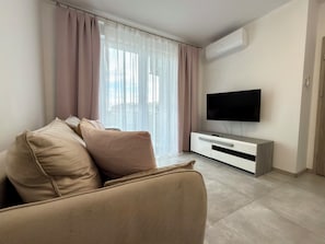 A modern living room with a beige sofa, a flat-screen TV on a white console, and a large window with sheer curtains. - Czartoryskiego Modern Apartment (Wrocław)