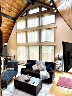 A cozy living room with a high ceiling, large windows, a sofa, a coffee table, and a dining area. - Majestic Hillside Escape (Anchorage)