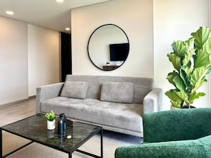 A modern living room with a grey sofa, a green armchair, a round mirror, and a large potted plant. - New Listing (Bogotá)
