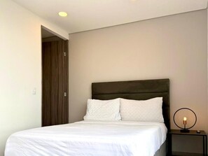 A neatly made bed with white bedding, a dark headboard, and a bedside table with a lamp. - New Listing (Bogotá)