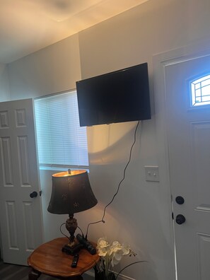 Ein Zimmer mit einem an der Wand befestigten Fernseher, einer Lampe auf einem kleinen Tisch und einer Tür mit einem Fenster.