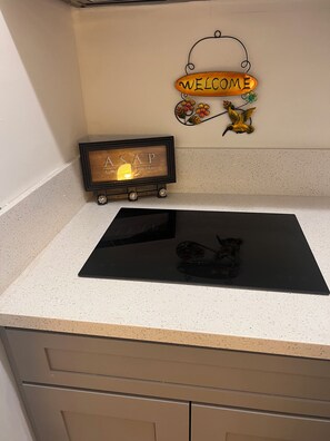 A kitchen counter with a black induction cooktop, a wooden sign with 'ASAP' and 'ALL WORKING PRAY' text, and a 'WELCOME' sign with yellow flowers. - Cozy 1-bedroom corporate apartment in Perris close Skidiving School and Hospital (Perris)