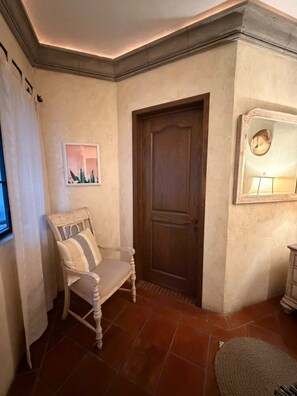 Um quarto com porta de madeira, uma cadeira com almofada listrada, um tapete pequeno e um espelho fixado na parede.