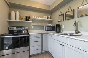 A compact kitchen with a stove, microwave, and sink. There are cabinets, a countertop, and wall-mounted shelves with various items. - Wall Street Suites 4 | Queen Studio near SIU (Carbondale)
