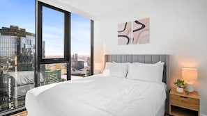 Ein Hotelzimmer mit einem großen Bett, einem Nachttisch mit Lampe und Blick über die Stadt durch ein großes Fenster.