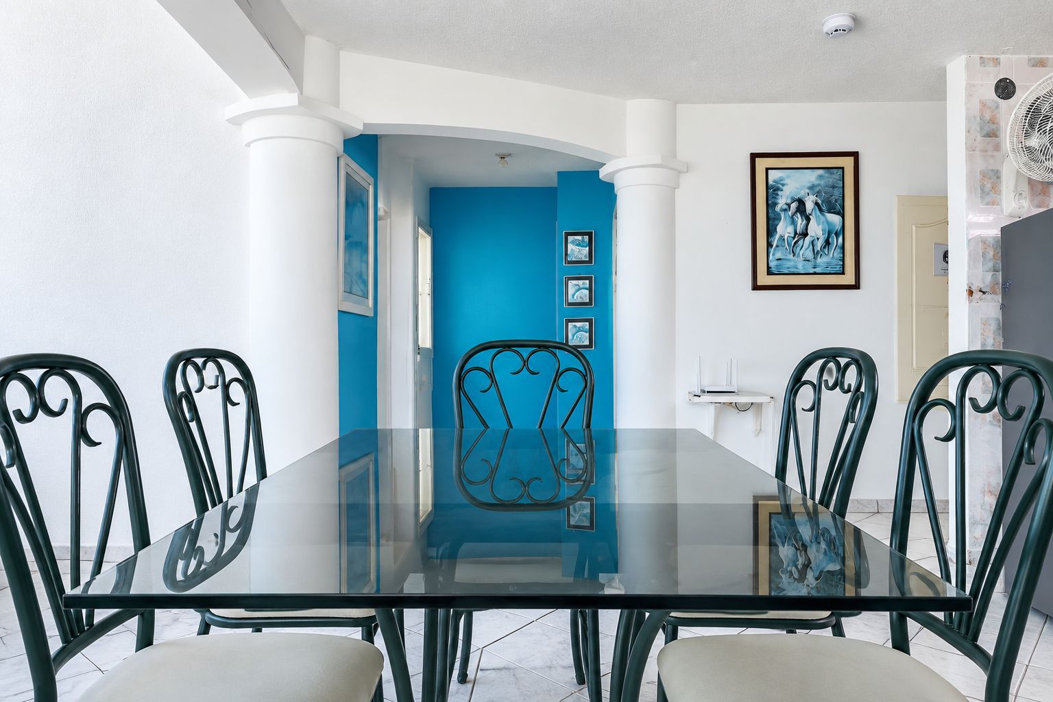 Un coin repas avec une table en verre et des chaises en métal, un mur d’accent bleu et des tableaux encadrés accrochés au mur.